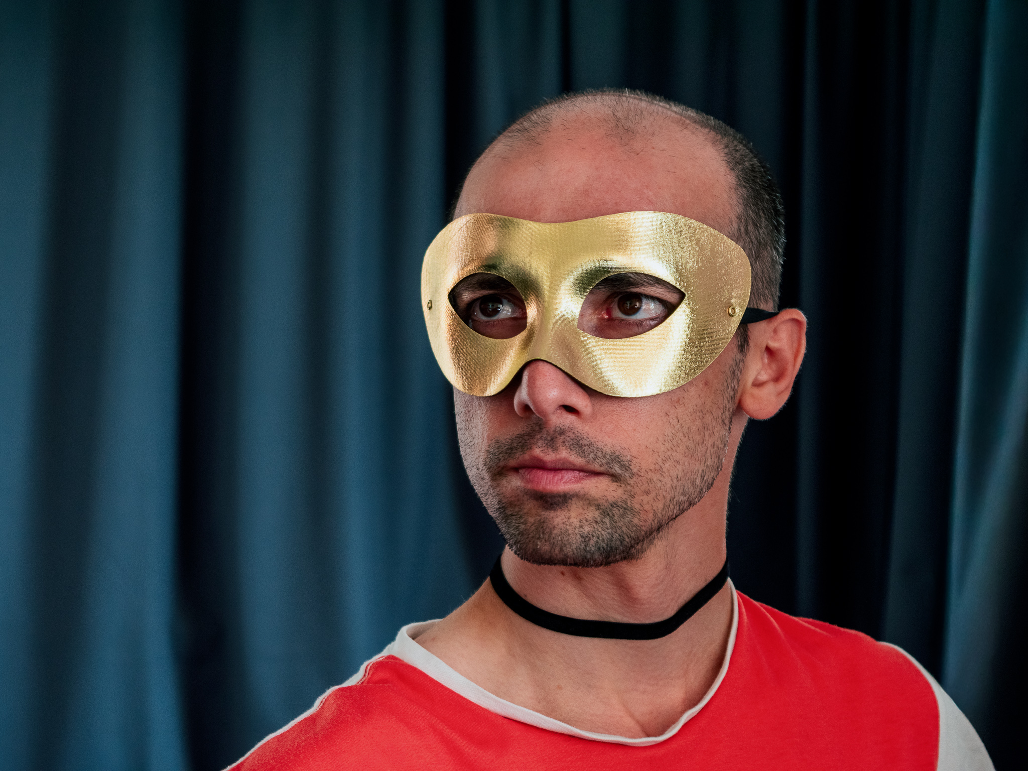 Close-up portrait of a man wearing a gold mask against dark curtains