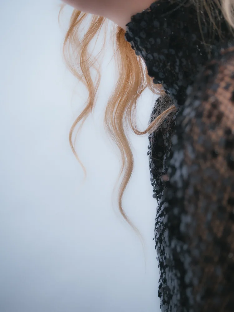 Fashion detail of wavy blonde hair beside a black sequined dress against a pale blue background