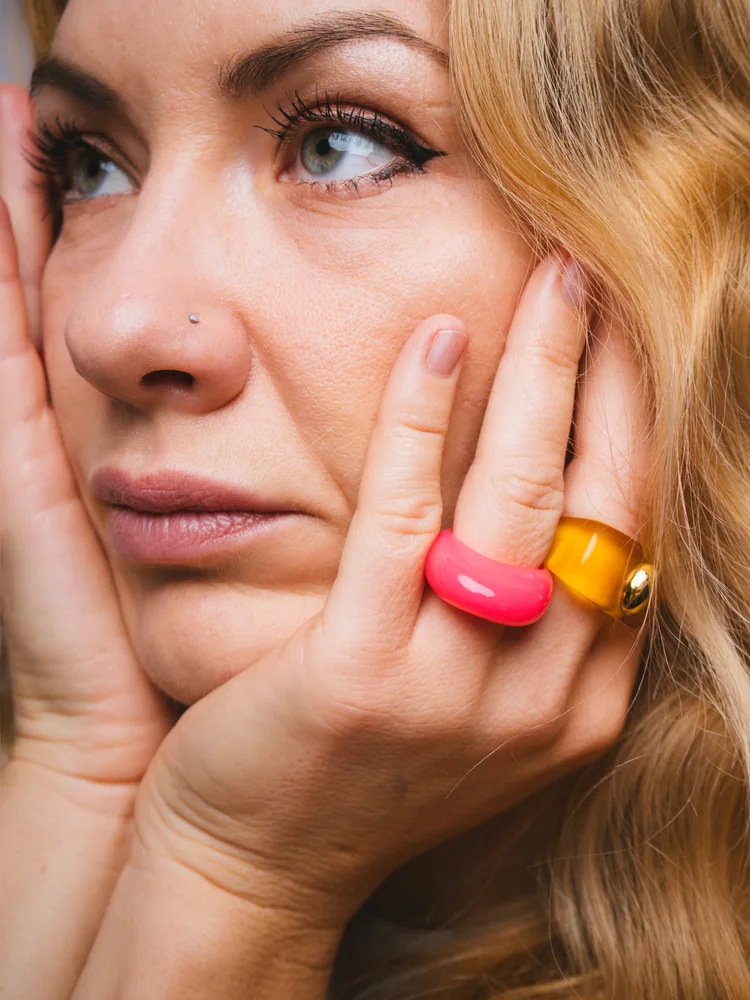 Close-up portrait of a woman resting her face in her hands, wearing bright pink and yellow rings