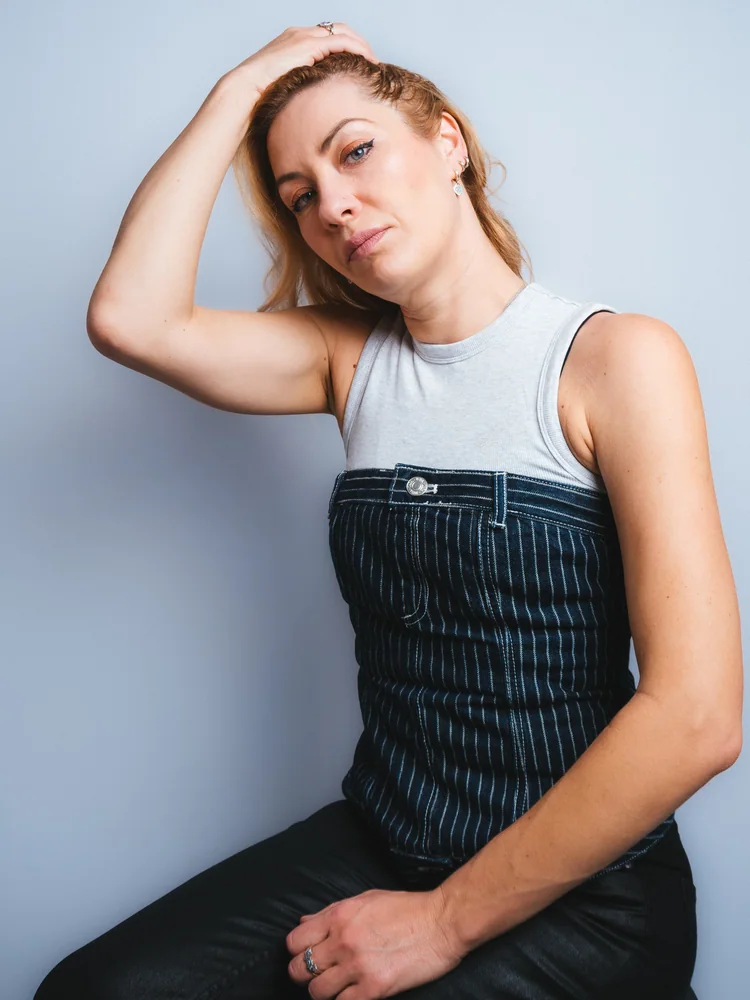 Studio portrait of a woman in a striped corset and white top, seated with one hand resting on her head
