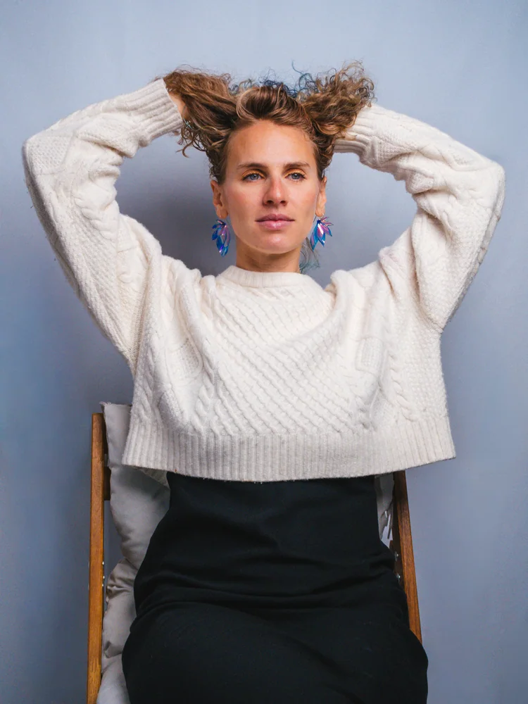 Studio portrait of a woman in a white sweater seated on a chair, lifting her curly hair with both hands