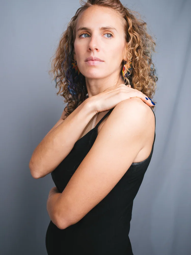 Studio portrait of a woman with curly hair and colorful earrings, one hand resting on her shoulder