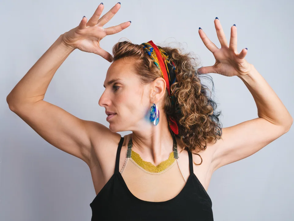 Portrait of a woman in a black top and colorful headband raising both hands beside her head