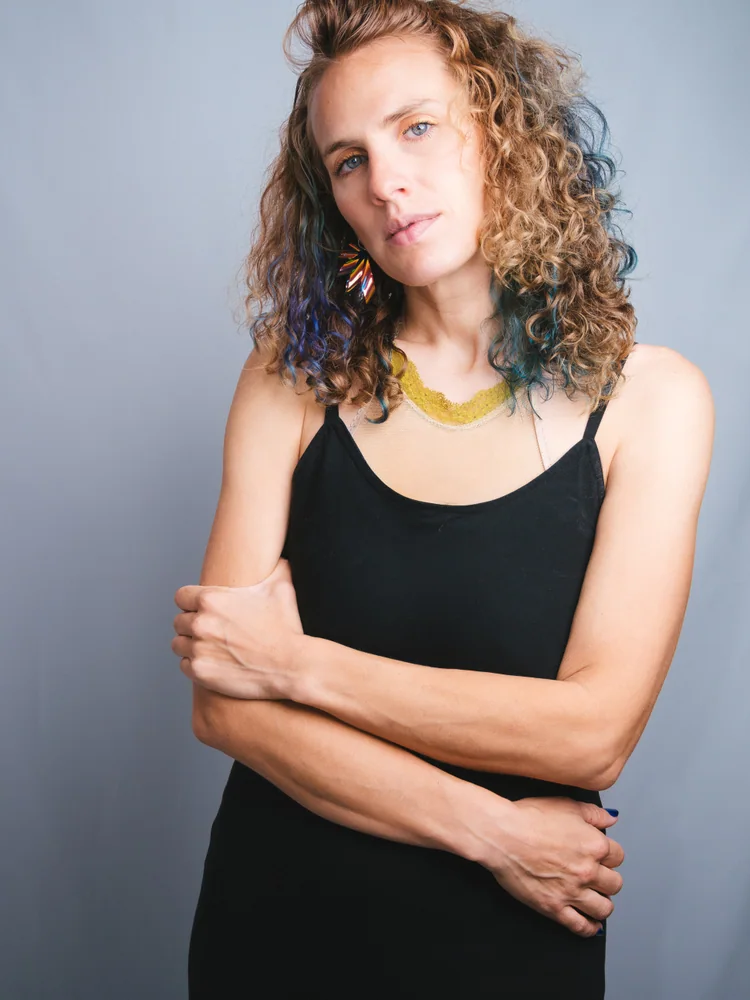 Portrait of a woman with curly hair and colorful earrings standing with her arms folded