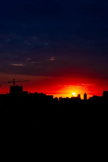 A bright setting sun above a silhouetted city skyline, with cranes beneath a deep blue and red sky.